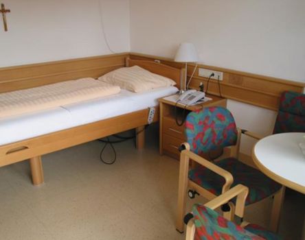 L'arredamento di una camera: un letto singolo regolabile in altezza, un comodino con telefono, un tavolino con due poltrone e una croce sulla parete accanto al letto