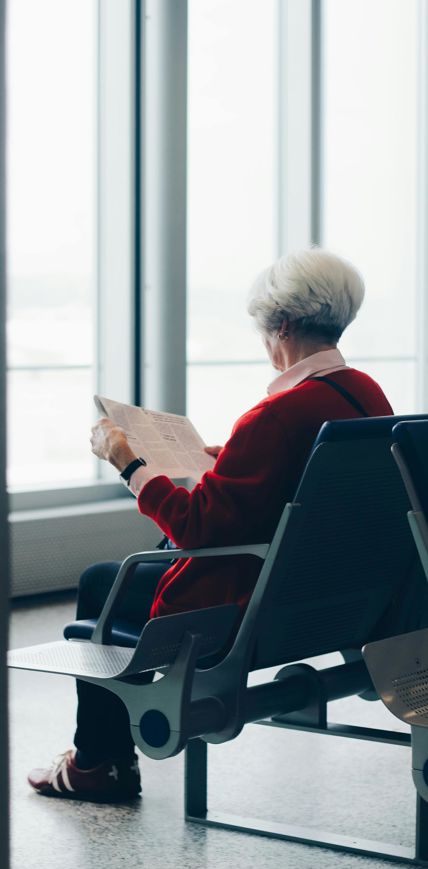 Eine ältere Frau mit weißem Haar und roter Jacke sitzt auf einem Stuhl in einem modernen Wartebereich und liest eine Zeitung, im Hintergrund große Fenster mit Tageslicht