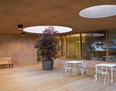 Moderna terrazza coperta con pavimento in legno, tavoli e sedie bianche, una panchina in legno e un albero in vaso che cresce attraverso un’apertura rotonda nel soffitto; ampie finestre riflettono l’esterno